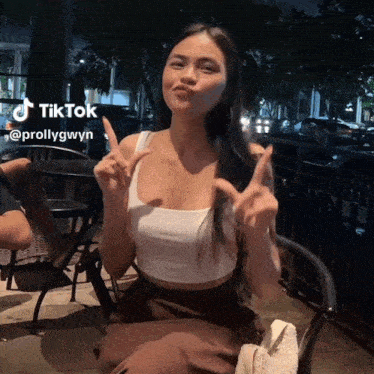 a woman in a white tank top is sitting in a chair and making a peace sign with her hands