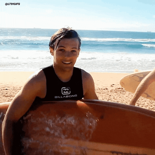 a man wearing a black billabong tank top holds a surfboard on the beach
