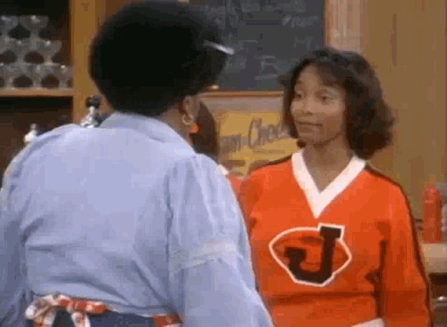 two women are standing next to each other in a kitchen . one of the women is wearing a red jersey with the letter j on it