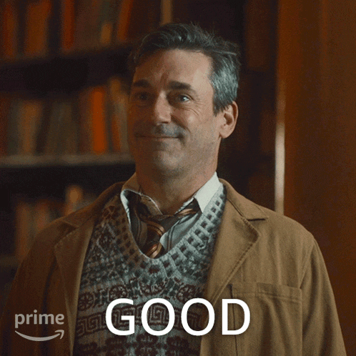 a man wearing a sweater and tie is smiling in front of a sign that says " good "