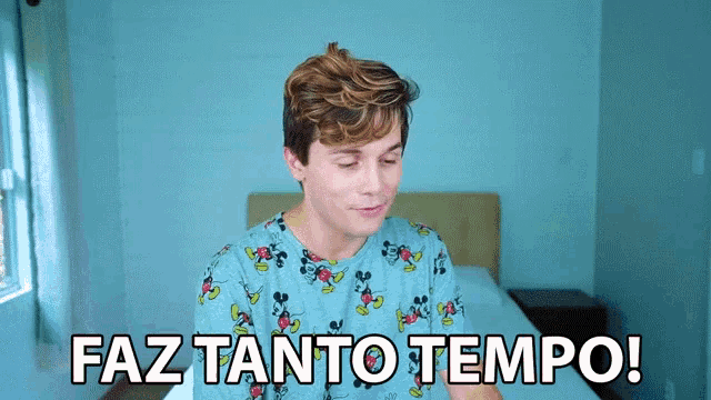 a young man wearing a mickey mouse shirt is sitting on a bed and says faz tanto tempo
