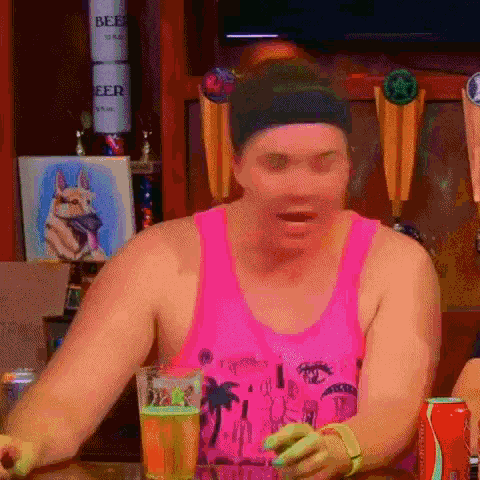 a man in a pink tank top is sitting at a table drinking beer .