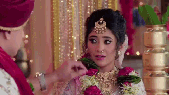 a bride and groom are standing next to each other and the bride is wearing a garland of flowers on her head .