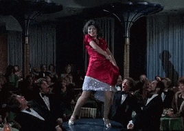 a woman in a red dress is dancing on a stage in front of a crowd of people in tuxedos .