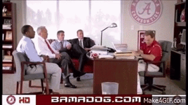a group of men are sitting around a table with a crimson tide logo on the wall