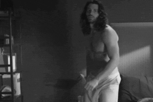 a shirtless man with long hair and a beard is standing in front of a bed