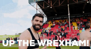 a man with a beard stands in front of a stadium with the words up the wrexham