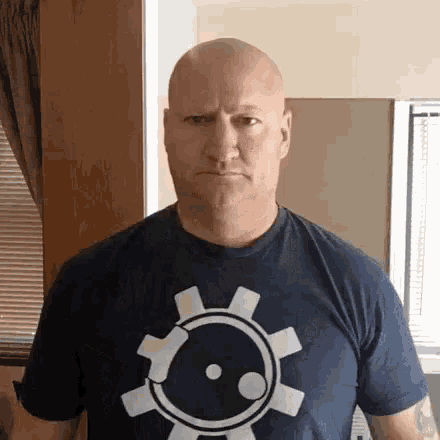 a bald man wearing a blue t-shirt with a gear on it is making an angry face .