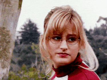 a blonde woman wearing glasses and a red turtleneck looks at the camera