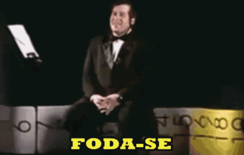 a man in a tuxedo sits in front of a microphone with the words foda-se written in yellow