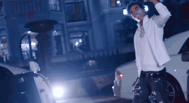 a man in a white jacket stands in front of a white car smoking a cigarette