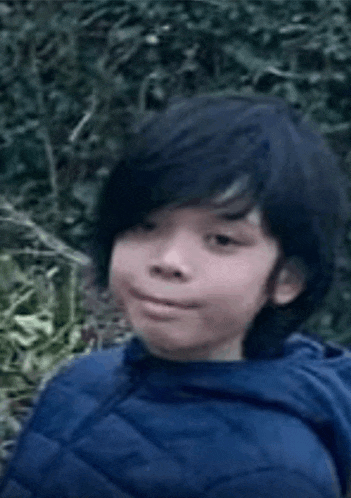a young boy with long black hair is wearing a blue jacket and making a funny face .