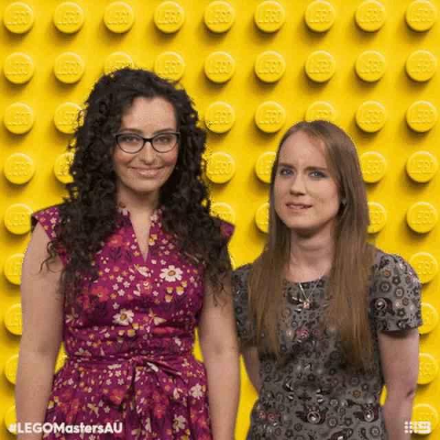 two women standing in front of a yellow wall with lego bricks on it