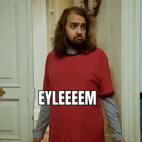 a man with long hair and a beard is wearing a red shirt that says eyleeeeem