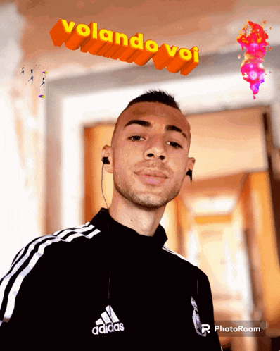 a man wearing a black adidas jacket stands in front of an orange sign that says volando voi