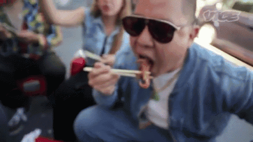 a man wearing sunglasses is eating food with chopsticks while sitting on the ground .