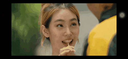 a woman is eating a candy with a toothpick in her mouth while looking at a man in a yellow jacket .