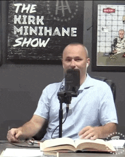 a man is sitting at a desk in front of a microphone and a sign that says the kirk minihane show