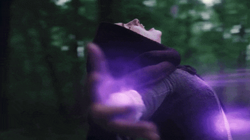 a woman in a black hood is standing in the woods with a purple light coming out of her mouth .