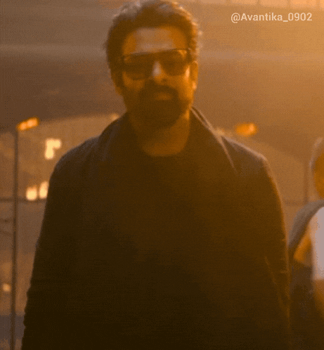 a man with a beard wearing sunglasses and a black jacket stands in a dark room