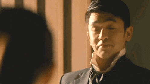 a man in a suit and tie looks at another man with his eyes closed