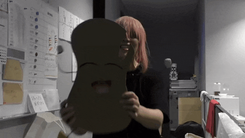 a woman with pink hair is holding a cardboard mask with a face on it