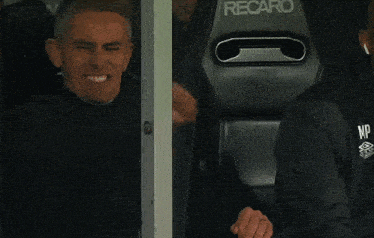 a man in a black jacket with np on the back is sitting in a recaro seat
