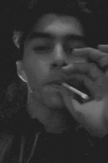a black and white photo of a young man smoking a cigarette in a dark room .