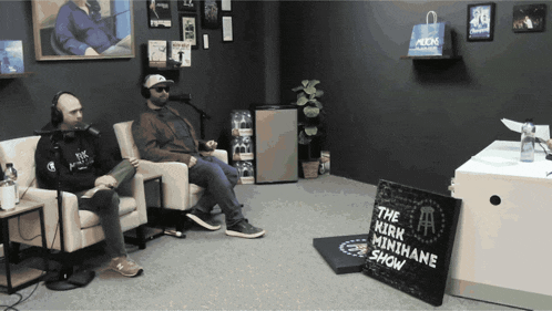 two men sit in chairs in a room with a sign that says the kirk minihane show