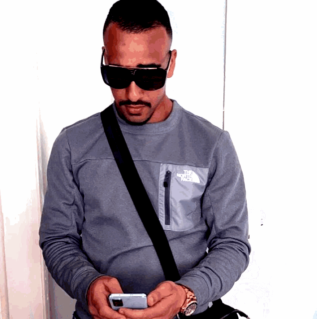 a man wearing sunglasses and a grey north face shirt looks at his phone