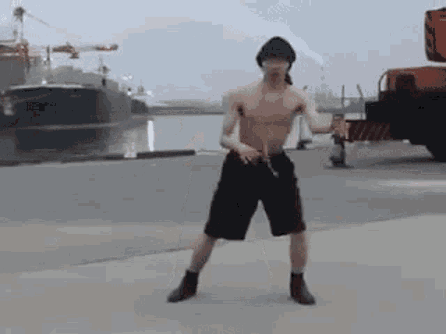 a man without a shirt is dancing in front of a truck and a boat .