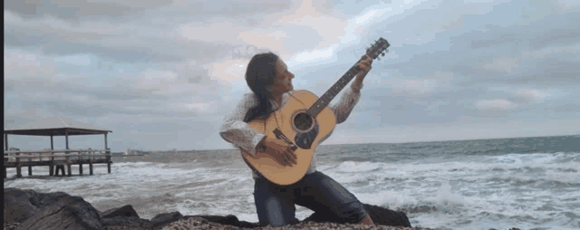 a woman kneeling on a rock holding an acoustic guitar in front of the ocean