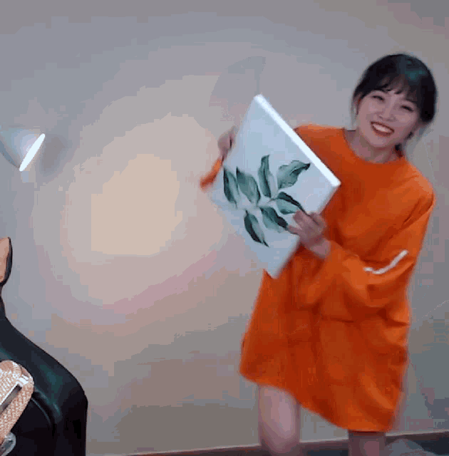 a woman in an orange dress is holding a white canvas with leaves on it