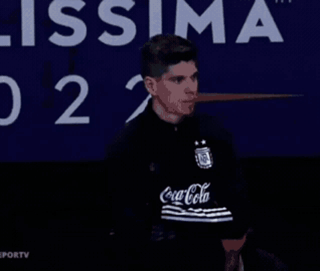 a man wearing a black coca cola shirt stands in front of a blue background