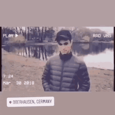 a man in a black jacket and hat is standing in front of a lake .
