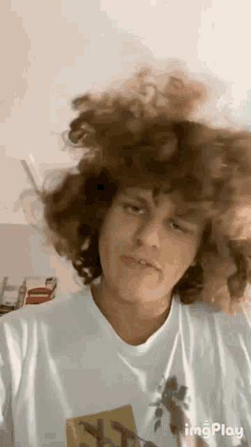 a young man with curly hair is wearing a white t-shirt and making a funny face .