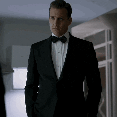 a man in a tuxedo and bow tie stands in a hallway with his hands in his pockets