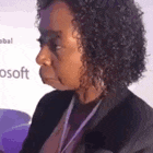 a woman with curly hair and a lanyard around her neck is sitting in front of a microsoft sign .