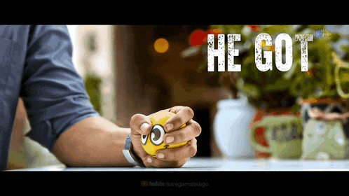 a man is holding a yellow ball with a face on it in front of a sign that says " he got "