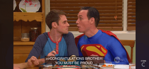 two men in superman costumes are sitting at a table and one of them is saying congratulations brother you must be proud