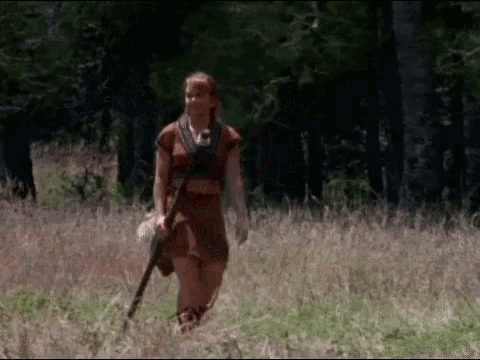 a woman walking through a field with a spear in her hand