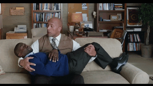 two men are laying on a couch and one of them has a blue shirt on