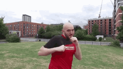 a bald man in a red shirt is standing in a grassy field in front of a building .