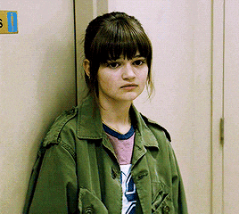 a woman in a green jacket leans against a wall with a sign on it that says ' s '