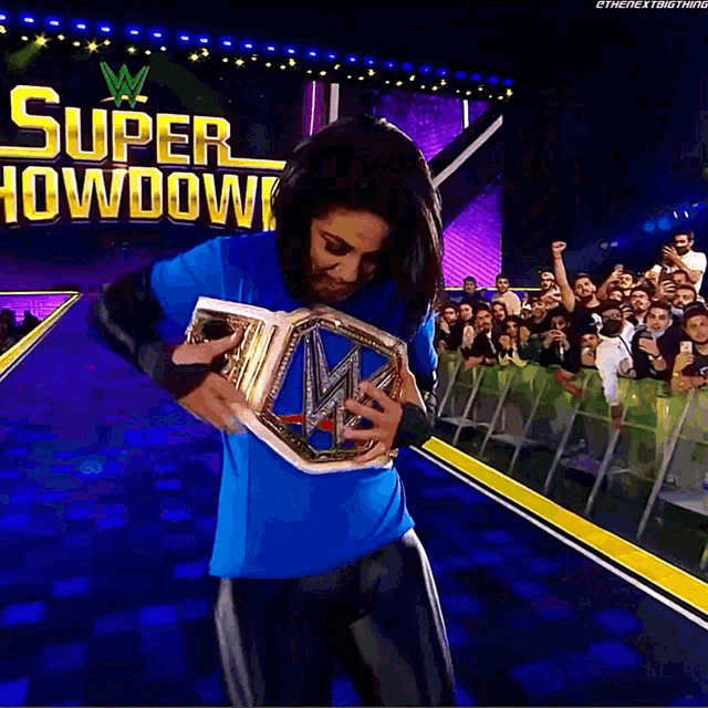 a woman in a blue shirt is holding a wrestling championship belt in front of a crowd at a super showdown