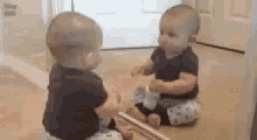 two babies are sitting on the floor looking at themselves in the mirror .