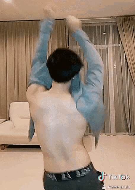 a man without a shirt is dancing in a living room with a tiktok logo on the bottom