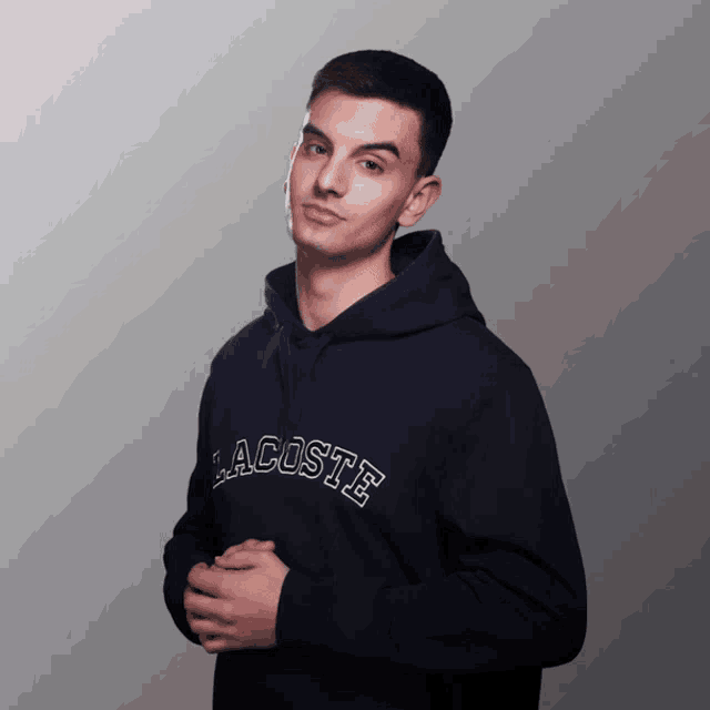 a young man wearing a black lacoste hoodie stands with his hands folded