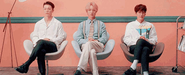 three men sit in chairs in front of a pink wall