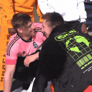 a man in a pink adidas shirt is hugging another man in a black jacket with a green barcode on the back